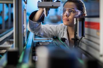 Jeune femme avec lunettes de protection examinant une pièce d'une machine de production