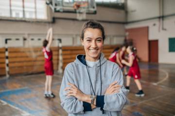 Une jeune fille souriante regardant l'objectif dans un gymnase, au fond des joueuses de baskets.